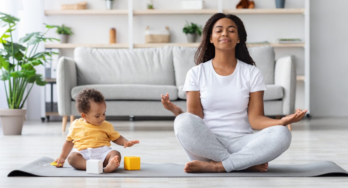 Mental Wellness Concept. Calm Happy Black Mother Meditating With Baby At Home, African Lady Sitting In Lotus Position With Closed Eyes, Practicing Yoga In Living Room With Child Playing Around