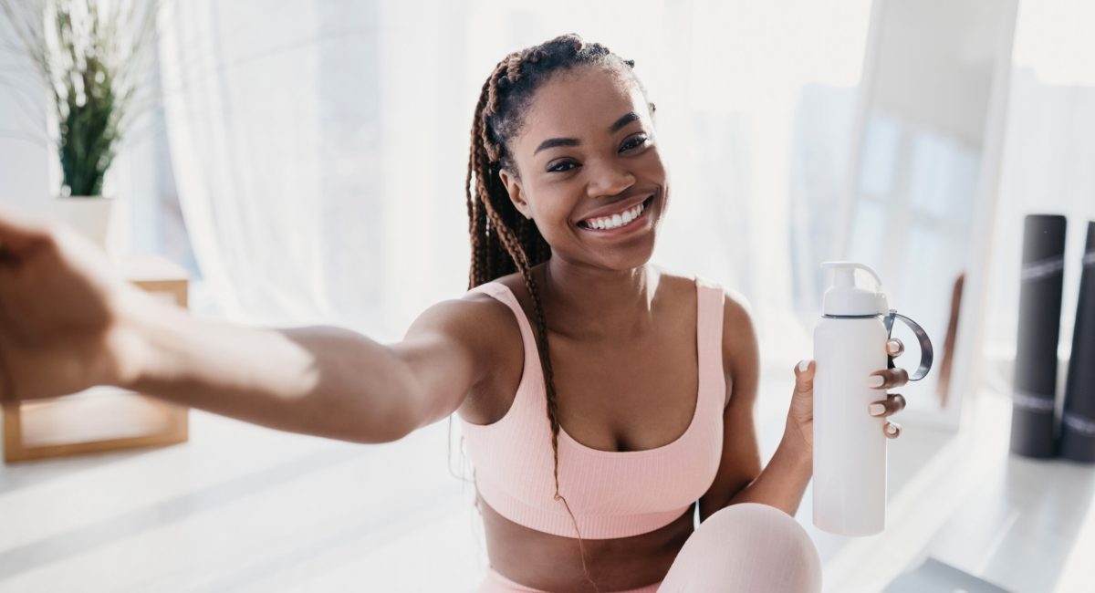 Smiling young African American lady with bottle of water taking selfie on break from home workout. Positive black woman making photo of herself during domestic training, indoors