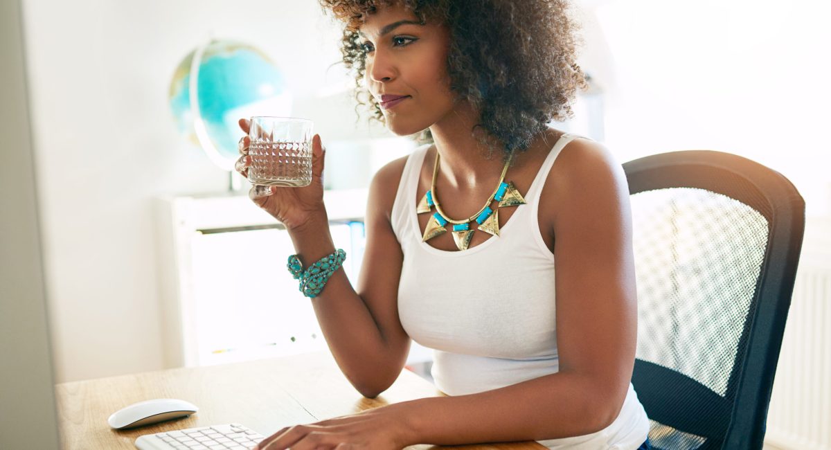 Young attractive African American businesswoman drinking water as she sits at her desk typing information on a desktop computer